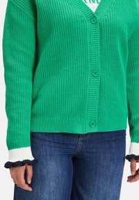 Zelený pletený cardigan se třemi knoflíky, nošený přes bílý vršek, v kombinaci s modrými džíny, s rukama po stranách.