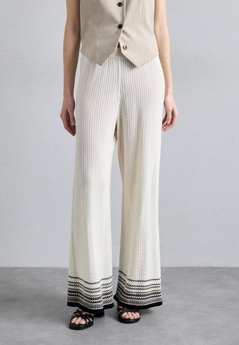 Crèmewitte, geribbelde wijde broek met een patroon op de zoom met zwarte lijnen. Gecombineerd met een beige, knoopvest en zwarte sandalen.
