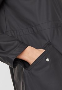 Czarna wodoodporna kurtka o gładkiej teksturze, z boczną kieszenią zabezpieczoną zatrzaskiem i subtelnymi detalami w szyciu.