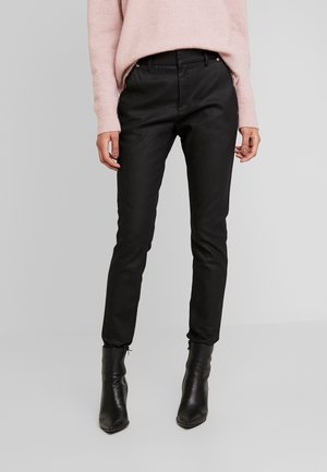 Frau trägt schwarze, eng anliegende Hose mit Knopf und Reißverschluss, hellrosa Pullover und schwarze Stiefeletten mit Absatz, steht vor einfachem Hintergrund.