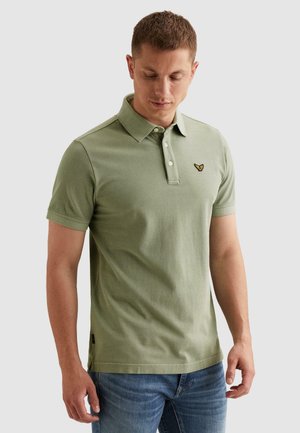 Muž v svetlozelenom krátkym rukávom polokošeli s golierom a malým vyšívaným logom na hrudi, oblečený v modrých džínsoch, stojaci na jednoduchom pozadí.