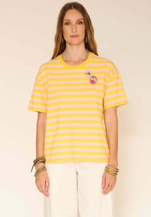 Femme portant un t-shirt rayé jaune et rose avec deux épingles rondes à thème de fruits, un pantalon blanc et plusieurs bracelets, debout devant un fond clair.