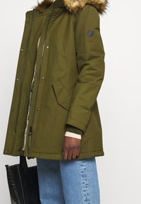 Olivengrøn parka med pelskrave, skrå forlommer og sort rund skulderlap, iført over lyseblå jeans og holder en sort taske.