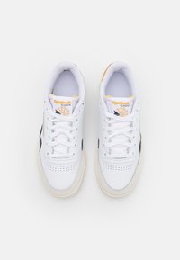 Sneaker Reebok Classic bianche con punta perforata, dettagli blu navy e logo giallo. Lacci in cotone e suola in gomma.