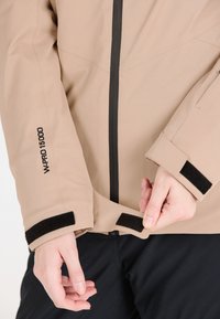 Beige jacka med en slät struktur. Har svart dragkedja, kardborrefästen och tryckt "W-PRO 15000" på ärmen. Närbild av justering vid ärmslut.