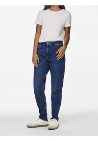 Perempuan berdiri mengenakan kaos putih lengan pendek, jeans biru pinggang tinggi, dan sepatu sneakers putih dengan sol krem.