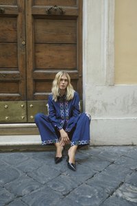 Mladá žena so svetlými vlasmi sedí na kamenných schodoch pred veľkými drevenými dverami, má na sebe modrý overal s vzorom a čierne špicaté lodičky.