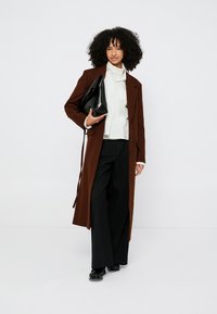 Cappotto lungo marrone in lana, indossato sopra una camicia bianca e pantaloni neri a gamba larga. Accessori comprendono una borsa nera. Riccioli texturizzati.