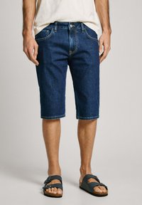 Donkere blauwe denimshorts met een rechte pasvorm, vijf zakken, beige stiksels en een knoopsluiting. Gecombineerd met donkere sandalen.
