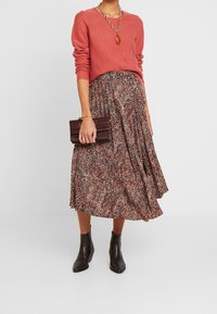 Czerwony sweter, spódnica midi w wzory w odcieniach brązu i czerwieni, czarne botki za kostkę oraz ciemnobrązowa kopertówka o teksturowanym wykończeniu.