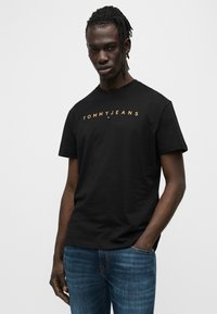 Μαύρο βαμβακερό t-shirt με το κείμενο "TOMMYJEANS" σε πορτοκαλί. Το σχέδιο έχει στρογγυλή λαιμόκοψη και κοντά μανίκια. Φοριέται με μπλε τζιν παντελόνι.