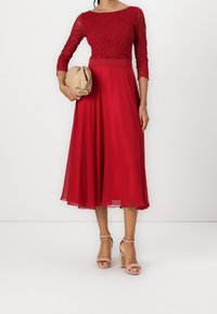 Robe à corsage en dentelle rouge avec des manches trois-quarts et une jupe fluide. Associée à une pochette beige et des talons nus à brides.