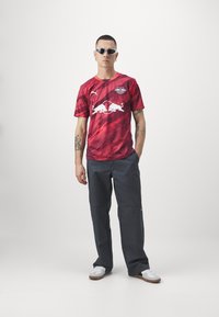 Maglia atletica a strisce rosse e nere con logo bianco e dettagli arancioni, abbinata a pantaloni grigi e sneakers bianche. Modello con occhiali da sole.