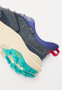 Scarpa da trekking con tomaia in rete blu e suede, collare viola, intersuola beige, suola turchese e logo Vibram giallo. Presenta occhielli in metallo.