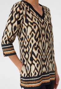 Langärmliges Oberteil in Creme, Schwarz und Orange mit V-Ausschnitt, das ein abstraktes Muster und kontrastierende Saumdetails aufweist.