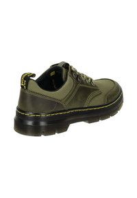 Olivgrüner Schuh mit Obermaterial aus texturiertem Canvas, verstärkter Zehenpartie und dicker schwarzer Gummisohle. Gelbe Nähte betonen die Sohle.