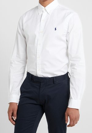 Homme portant une chemise blanche ajustée à boutons avec des manches longues et un pantalon bleu marine, debout devant un fond uni.