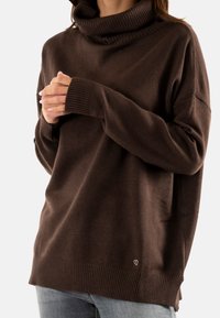 Maglione oversize marrone con collo a imbuto e polsini a costine. Presenta una texture liscia e un piccolo logo sull'orlo.