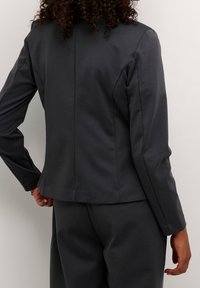 Chaqueta gris ajustada con mangas largas, cuello redondo y textura suave. Presenta una forma entallada y un dobladillo ligeramente corto en la parte posterior.