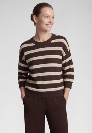 Woman with brown hair in a bun wears a brown and beige striped sweater and brown pants, smiling and looking to the right with hands in pockets.