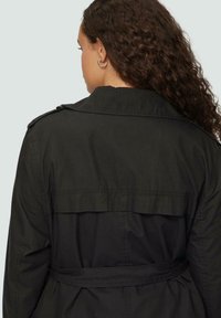 Schwarzer Trenchcoat mit tailliertem Gürtel, doppellagigem Rücken, Schulterklappen und einer glatten, matten Textur.