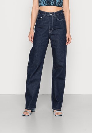 Woman wearing high-waisted dark blue straight-leg jeans, a cropped blue patterned top, and light blue open-toe heels standing against a white background.