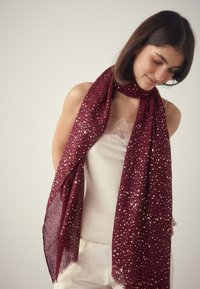 Burgunderroter Schal mit goldenen Sprenkeln, leichter Textur, lang und fließend, um den Hals getragen. Glatte Oberfläche, zarte Fransenränder.