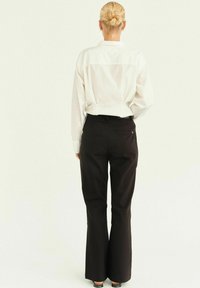 Pantaloni neri svasati con una texture liscia, dotati di vita aderente e tasche posteriori, abbinati a una camicia bianca con colletto dal taglio accattivante.