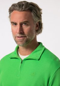 Pullover verde con zip realizzato in tessuto a coste e colletto alto. Presenta un piccolo logo sul petto e una t-shirt bianca sottostante.