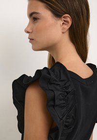 Haut noir avec des manches volantes et un col rond. Le tissu a une texture lisse, avec des détails froncés le long des manches.