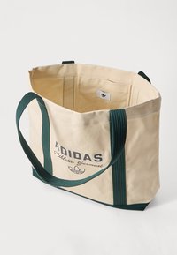Bežs Adidas tote somiņa ar tumši zaļām rokturiem un apmalēm, uz priekšpuses ir uzraksts un logo "Adidas Athletic Garment", atvērta augšdaļa, kurā redzama iekšējā birka.