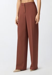 Pantalones de pierna ancha en un profundo color terracota, hechos de una tela suave. Presentan una cremallera frontal y un diseño minimalista. Combinados con zapatos beige de punta.