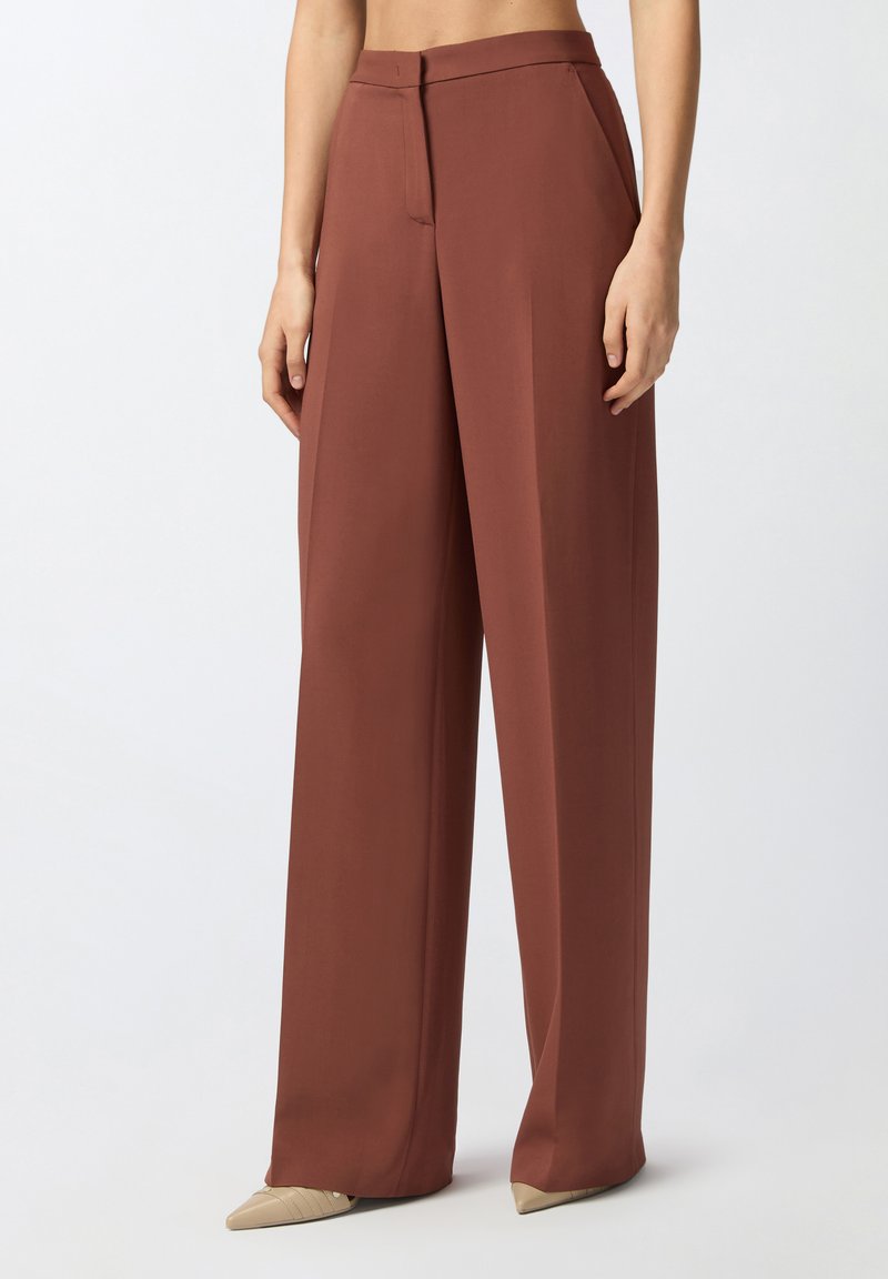 Pantalones de pierna ancha en un profundo color terracota, hechos de una tela suave. Presentan una cremallera frontal y un diseño minimalista. Combinados con zapatos beige de punta.