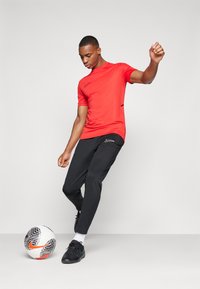 Rød atletisk T-skjorte med kort erm og svart Nike-logo. Svart treningsbukser med sidelommer. Svart sko og en fotball ved føttene.