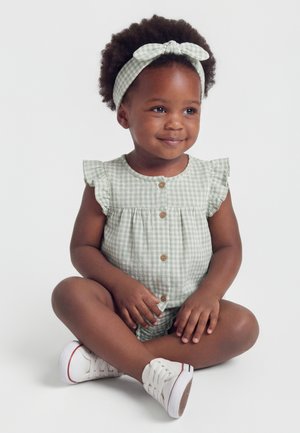 Fetiță mică cu păr creț, purtând un romper verde în carouri, bentiță asortată și adidași albi, zâmbind și stând așezată cu picioarele încrucișate.