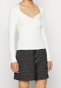 Haut en maille blanc à manches longues avec un design torsadé à l'avant, accompagné d'une jupe en tweed noire avec boutons sur le devant et accents brillants.