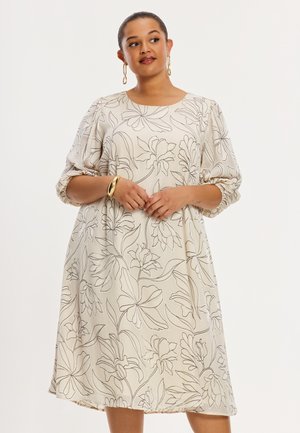 Vrouw draagt een crèmekleurige jurk tot op de knieën met zwarte bloemmotieven in lijnpatroon, driekwart pofmouwen, gouden oorringen en een gouden armband.