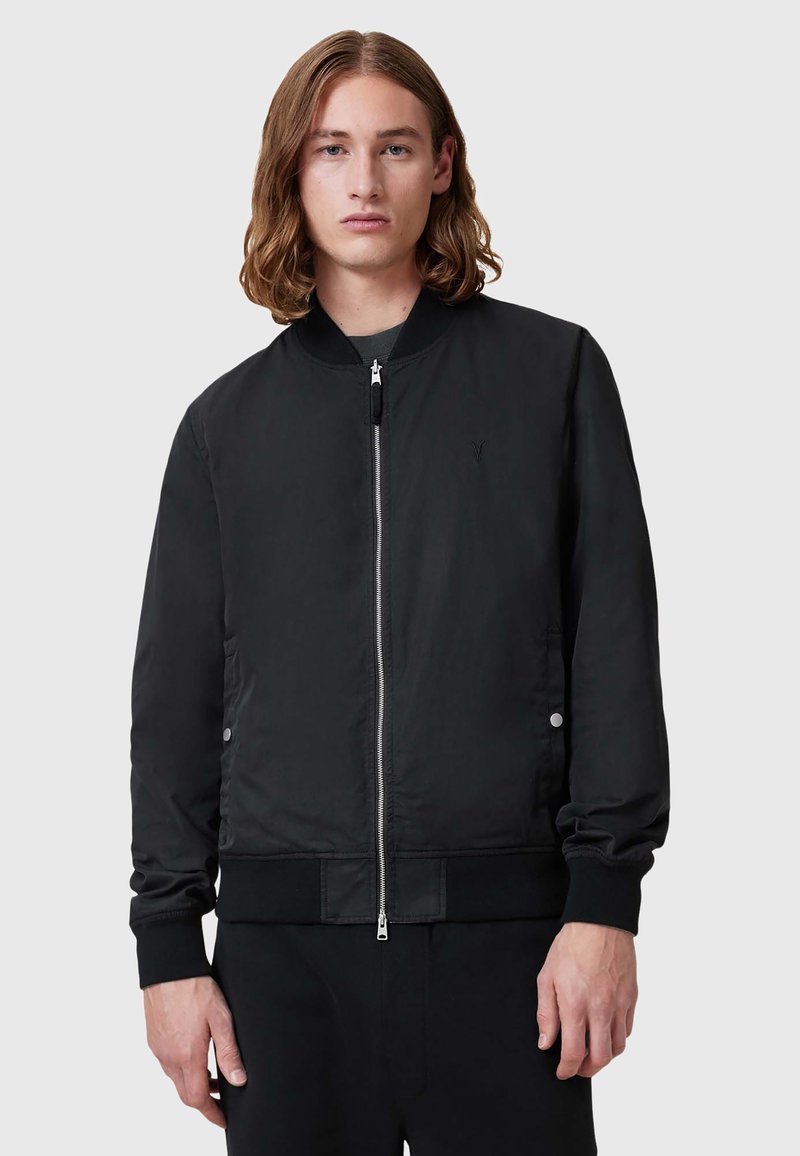 Schwarze Bomberjacke aus glattem Material, mit durchgehendem Reißverschluss, gerippten Bündchen und einem dezenten Logo auf der Brust. Schlichtes Design und Struktur.