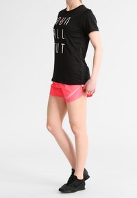 Camiseta atlética negra con gran texto blanco y pantalones cortos de correr rosa. Los pantalones cortos tienen una capa transparente y se combinan con zapatillas negras.