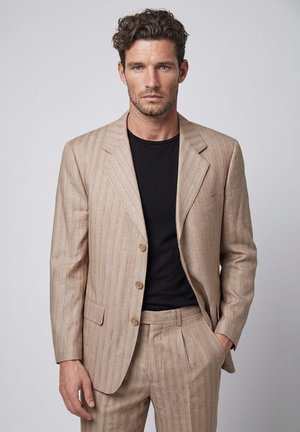 Hombre con cabello rizado que lleva un traje beige de rayas y camisa negra, de pie con una mano en el bolsillo contra un fondo liso y claro.