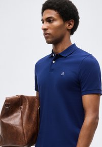 Άνδρας με μπλε πόλο πουκάμισο κρατώντας μια μεγάλη καφέ δερμάτινη τσάντα μπροστά σε ένα απλό φωτεινό φόντο.