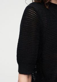 Haut court à manches courtes en crochet noir avec un bord-côte porté par une personne sur un fond clair uni.