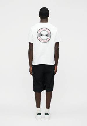 Άνδρας που φοράει λευκό μπλουζάκι Michael Kors με μαύρο σορτς και λευκά αθλητικά παπούτσια, στέκεται με την πλάτη σε απλό φόντο.