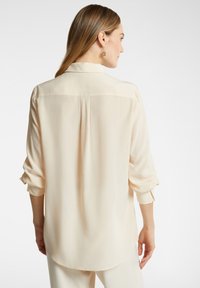 Crème-kleurige blouse met lange mouwen, gemaakt van lichte stof en met een ontspannen pasvorm, voorzien van een rugnaad en opgerolde mouwmanchetten.