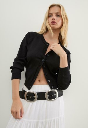 Schwarze gestrickte Strickjacke mit rundem Ausschnitt und Knopfverschluss. Kombiniert mit einem weißen Rock und einem breiten schwarzen Gürtel mit großen silbernen Schnallen.