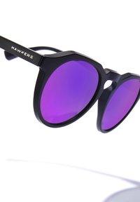 Gafas de sol con montura negra mate y grandes lentes redondos teñidos de púrpura. Presentan una forma hexagonal y un pequeño recorte en el puente.