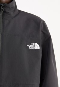 Chaqueta negra con cremallera hecha de tela lisa. Cuenta con un cuello alto y un logo blanco prominente en el área del pecho izquierdo.