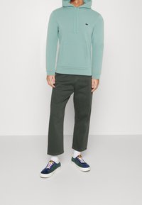 Felpa verde menta con tasca a kangaroo, abbinata a pantaloni larghi verde scuro e sneakers navy con suole bianche e lacci colorati.