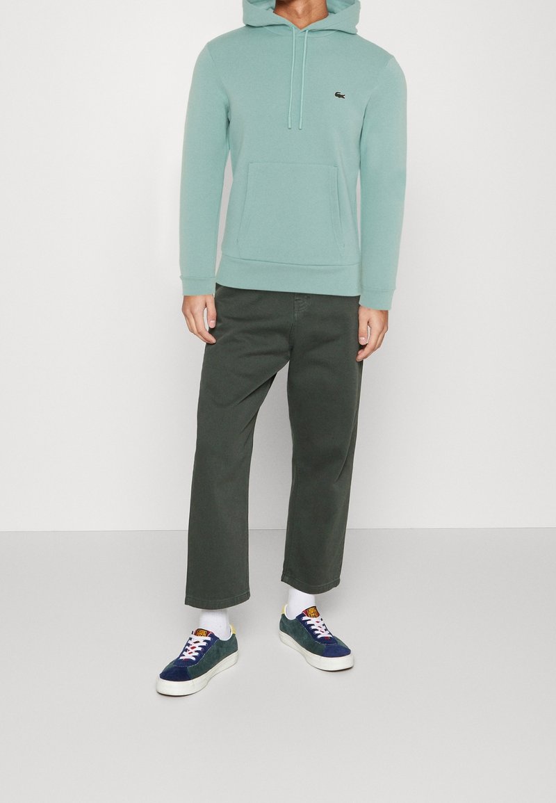Felpa verde menta con tasca a kangaroo, abbinata a pantaloni larghi verde scuro e sneakers navy con suole bianche e lacci colorati.