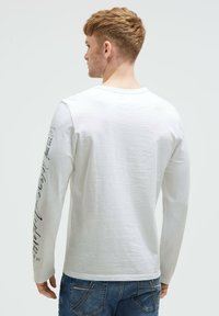 White long-sleeve shirt with a crew neck and printed text on the left sleeve. Worn with blue denim jeans, showing a casual fit.